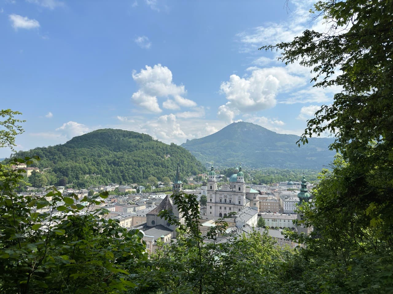 Historic city with green-domed cathedral towers nestled beneath forested hills, seen from a tree-framed overlook