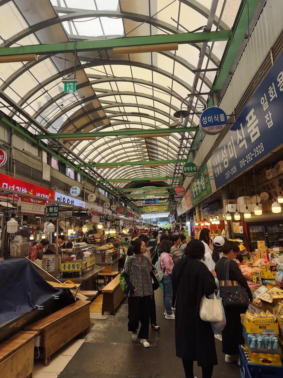 Shoppers strolling beneath the arched glass roof of a covered Korean market lined with food stalls and hanging signs