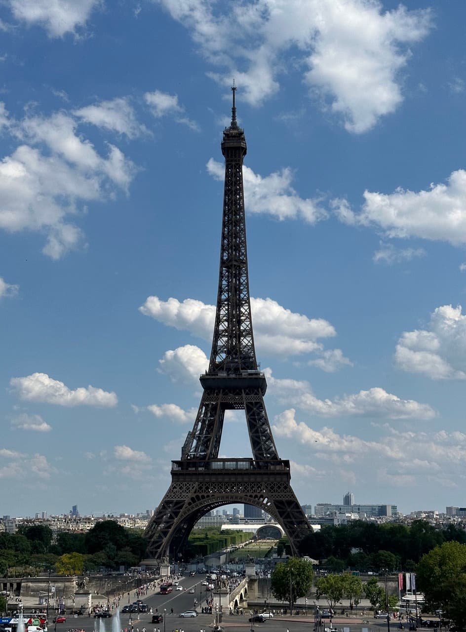 The Eiffel Tower rising into a blue sky dotted with clouds, with city rooftops and a busy street below