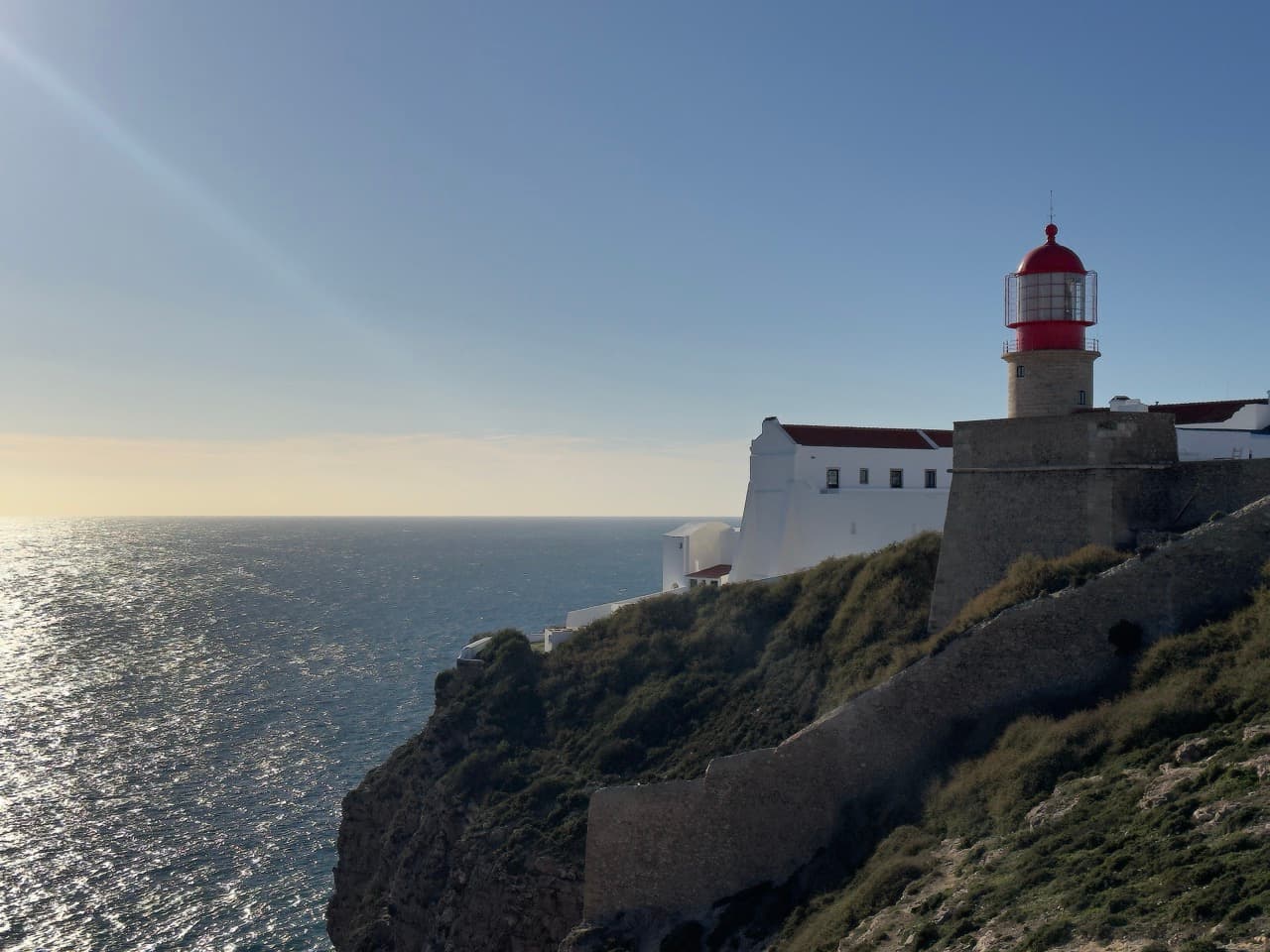 A red-capped lighthouse on stone fortifications at the edge of a sea cliff, sunlight shimmering on the ocean