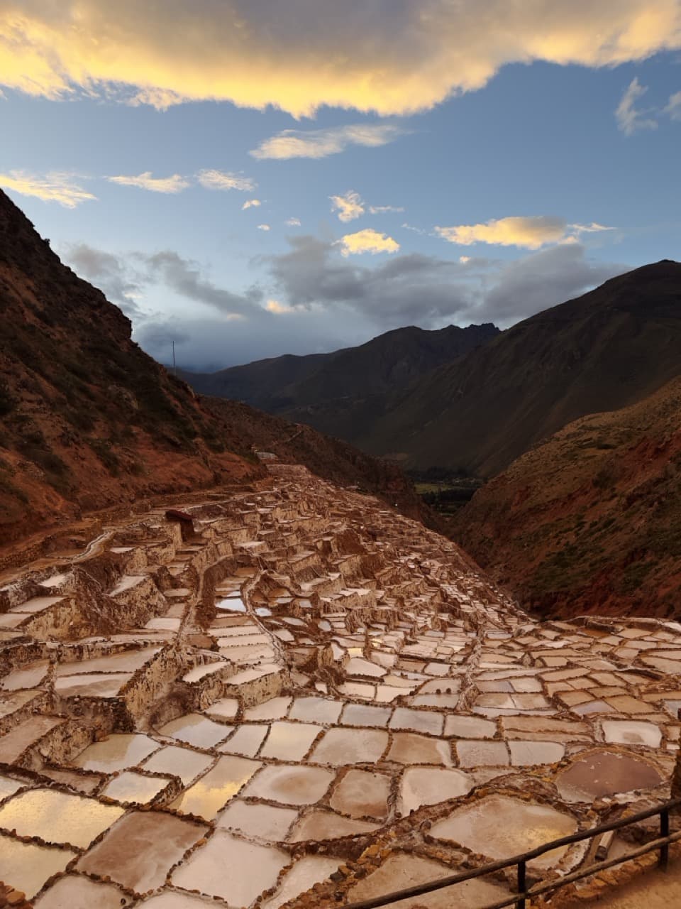 Terraced salt evaporation ponds cascading down a mountain valley under a golden sunset sky